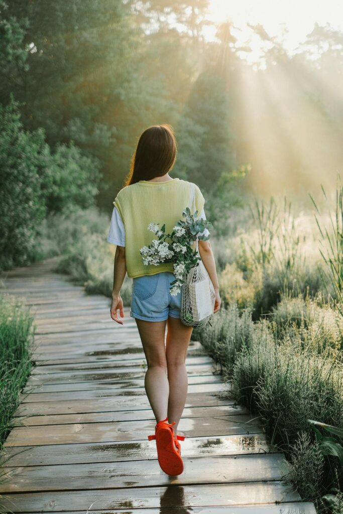 A woman walks along a sunlit wooden path in a serene forest setting, carrying flowers in a bag.