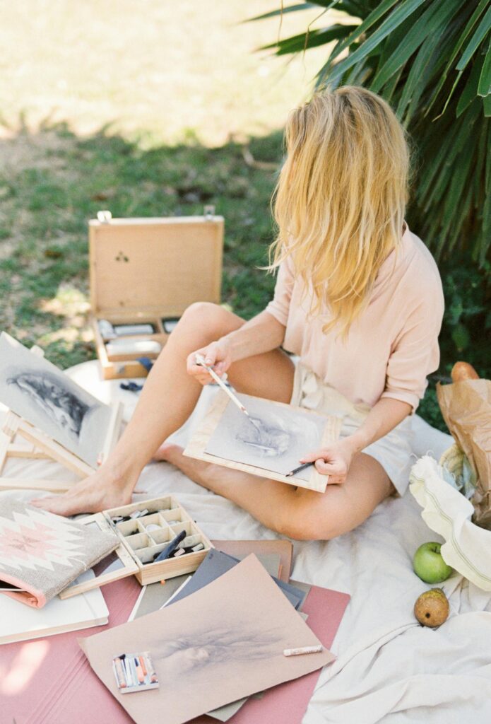 Blonde woman sketching on canvas outdoors, surrounded by art materials on a picnic blanket.