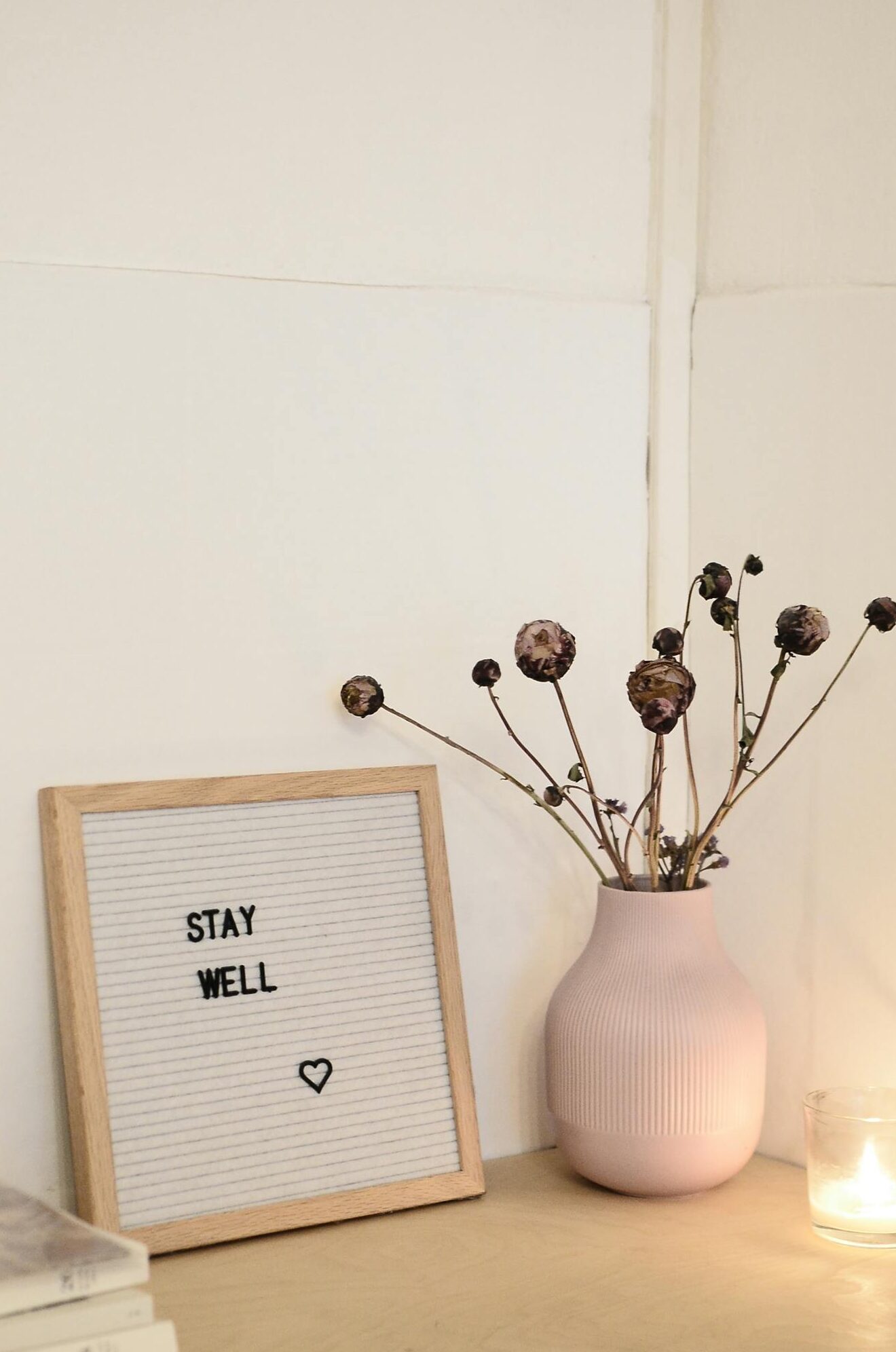 Corner of cozy room decorated with dry flowers in vase near frame with words stay well and stack of magazines