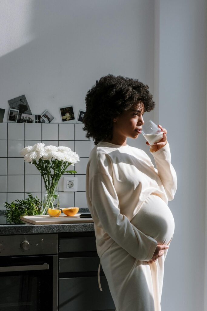 Pregnant woman drinking milk in a sunlit kitchen, surrounded by flowers and fruit.